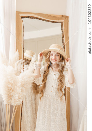 A beautiful teenage girl with long hair measures a straw hat in front of a mirror. Self-admiration of a blonde. 104802820