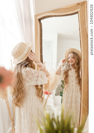 A beautiful teenage girl with long hair measures a straw hat in front of a mirror. Self-admiration of a blonde. 104802826