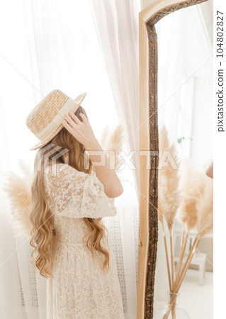 A beautiful teenage girl with long hair measures a straw hat in front of a mirror. Self-admiration of a blonde. A beautiful teenage girl with long hair measures a straw hat in front of a mirror. Self-admiration of a blonde. 104802827