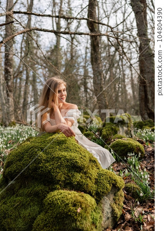 Snowdrops galanthus blonde. A girl in a white dress sits on a meadow with snowdrops in a spring forest 104804390