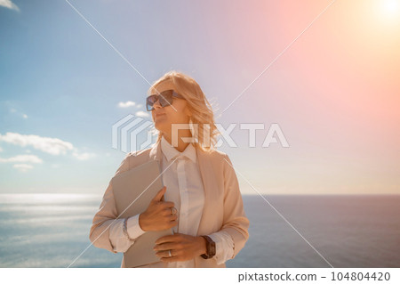 Freelance women sea working on a computer. Pretty middle aged woman with computer and phone outdoors with beautiful sea view. The concept of remote work. Freelance women sea working on a computer. Pretty middle aged woman with computer and phone outdoors with beautiful sea view. The concept of remote work. 104804420