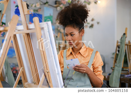 Happy young woman with Afro haired wearing apron painting picture with brush on canvas in art studio 104805158