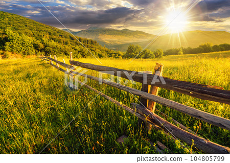 rural landscape at sunset. wooden fence near the grassy meadow and trees on the hillside. beautiful countryside scenery in evening light 104805799