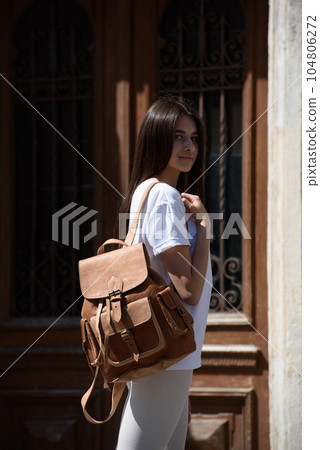 beautiful brunette girl in white leggings and T-shirt posing with a beige leather backpack. 104806272