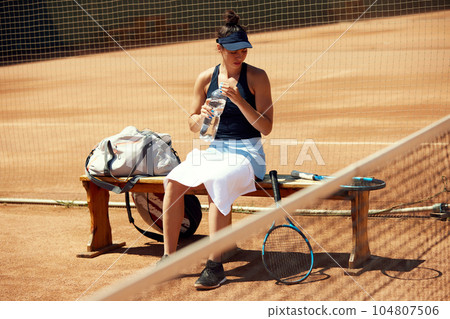 Young sportive girl in sportswear sitting on bench on open air tennis court after training, game resting and drinkign water 104807506