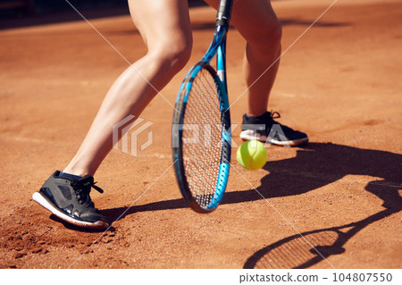 Cropped image of female legs in sportswear and tennis racket with ball over floor background with shadows. Open air tennis court 104807550