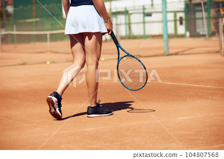 Cropped image of female legs in sportswear and tennis racket at open air tennis court on sunny warm day Cropped image of female legs in sportswear and tennis racket at open air tennis court on sunny warm day 104807568