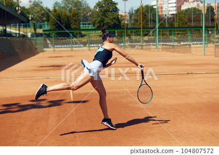 Concentrated female tennis player in motion during game, raining, playing tennis at open air court on sunny day Concentrated female tennis player in motion during game, raining, playing tennis at open air court on sunny day 104807572