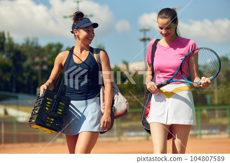 Young girls in sportswear going to play tennis on warm sunny day at open air tennis court. Friends meeting Young girls in sportswear going to play tennis on warm sunny day at open air tennis court. Friends meeting 104807589