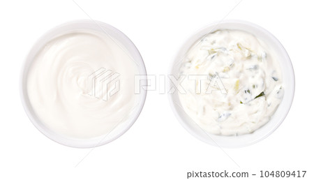 Cream yogurt and tzatziki, in white bowls. Stirred cream yoghurt with high fat content. Pure (left side), and as Greek dip sauce, mixed with cucumber, garlic, salt, olive oil and herbs (right side). 104809417