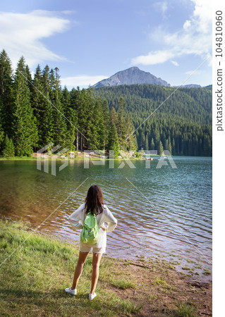 A girl with a bright backpack stands with her back near a mountain lake in the background. Positive young woman traveling on a blue lake outdoors. The girl admires the view. 104810960