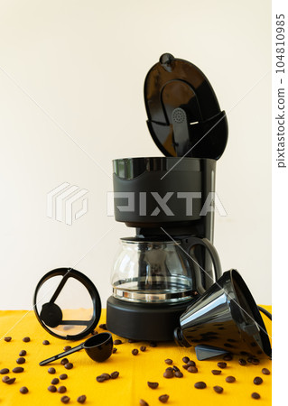 Drip black coffee maker Home appliances on the kitchen table. Preparation of hot coffee, scattered grains on a bright napkin. Drip black coffee maker Home appliances on the kitchen table. Preparation of hot coffee, scattered grains on a bright napkin. 104810985