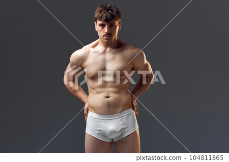Handsome young man with muscular body, relief torso. Model posing shirtless in underwear against grey studio background Handsome young man with muscular body, relief torso. Model posing shirtless in underwear against grey studio background 104811865