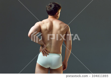 Pain in spine. Cropped image of muscular male body, relief back. Man posing shirtless in underwear against grey studio background Pain in spine. Cropped image of muscular male body, relief back. Man posing shirtless in underwear against grey studio background 104811893