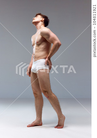 Full-length image of young man with healthy, strong, muscular body posing shirtless in underwear against grey studio background. Back pains Full-length image of young man with healthy, strong, muscular body posing shirtless in underwear against grey studio background. Back pains 104811921