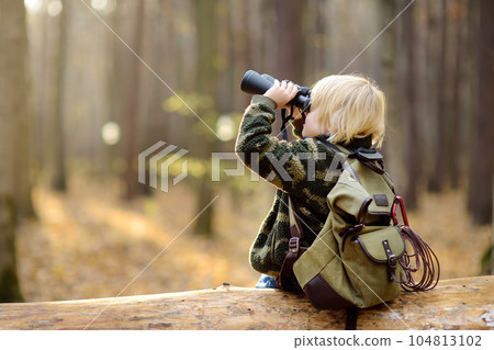 Little boy scout with binoculars during hiking in autumn forest. Child is sitting on large fallen tree and looking through a binoculars. Little boy scout with binoculars during hiking in autumn forest. Child is sitting on large fallen tree and looking through a binoculars. 104813102