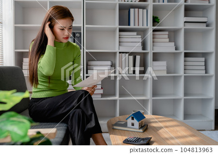 woman showing an anxious expression sitting on sofa and planning and calculate expense and mortgage with calculator and home on desk, insurance and budget of residential, loan and residence 104813963