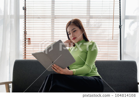 Asian young woman making notes in paper notebook sitting on cozy sofa on background of window. Pretty female writing notes in diary or making to do list relaxing at home Asian young woman making notes in paper notebook sitting on cozy sofa on background of window. Pretty female writing notes in diary or making to do list relaxing at home 104814108