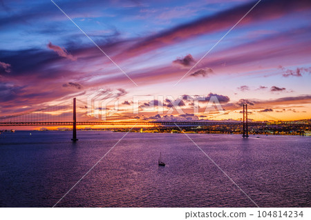 View of 25 de Abril Bridge famous tourist landmark of Lisbon connecting Lisboa and Almada on Setubal Peninsula over Tagus river in the evening twilight with boats. Lisbon, Portugal View of 25 de Abril Bridge famous tourist landmark of Lisbon connecting Lisboa and Almada on Setubal Peninsula over Tagus river in the evening twilight with boats. Lisbon, Portugal 104814234
