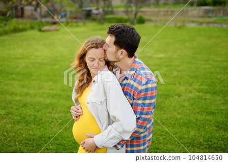 Young family is expecting a baby. Happy couple of parents-to-be walking together. Husband embracing wife gently and holding her belly enjoying. 104814650