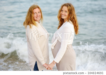 Redhead senior mother and her adult beautiful pregnant daughter are walking together on the sea shore. A happy meeting of a mom and her grown child. Redhead senior mother and her adult beautiful pregnant daughter are walking together on the sea shore. A happy meeting of a mom and her grown child. 104814691