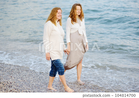 Redhead senior mother and her adult beautiful pregnant daughter are walking together on the sea shore. 104814700