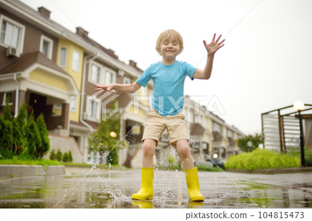 Little boy wearing yellow rubber boots jumping in puddle of water on rainy summer day in small town. Child having fun. Outdoors games for children in rain. Little boy wearing yellow rubber boots jumping in puddle of water on rainy summer day in small town. Child having fun. Outdoors games for children in rain. 104815473