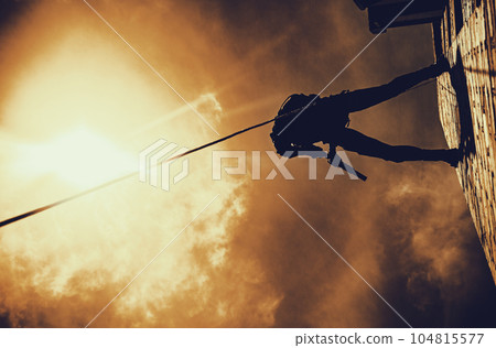 Silhouette of police officer in tactical gear descending from a height, rope exercises with weapons. Tactical rappelling, anti-terror or counter terrorism operation in darkness in rappelling harness Silhouette of police officer in tactical gear descending from a height, rope exercises with weapons. Tactical rappelling, anti-terror or counter terrorism operation in darkness in rappelling harness 104815577