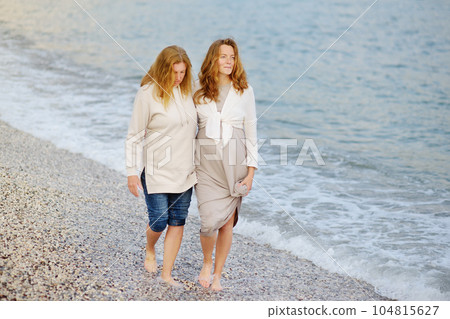 Redhead senior mother and her adult beautiful pregnant daughter are walking together on the sea shore. 104815627