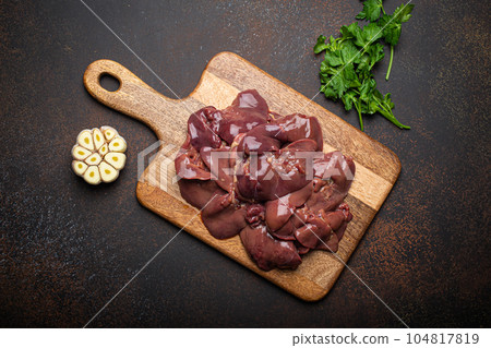 Raw chicken liver on wooden cutting board top view on dark rustic concrete background kitchen table with parsley and garlic. Healthy food ingredient, source of iron, folate, vitamins, minerals Raw chicken liver on wooden cutting board top view on dark rustic concrete background kitchen table with parsley and garlic. Healthy food ingredient, source of iron, folate, vitamins, minerals 104817819