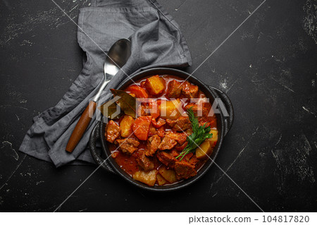 Beef meat stew with potatoes, carrot and delicious gravy in black casserole pot with bay leaves with spoon on black dark rustic concrete background from above . 104817820