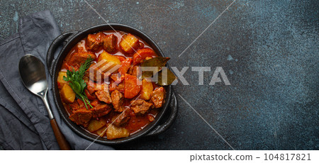 Beef meat stew with potatoes, carrot and delicious gravy in black casserole pot with bay leaves and fresh green herbs with spoon on dark rustic concrete background from above, space for text. Beef meat stew with potatoes, carrot and delicious gravy in black casserole pot with bay leaves and fresh green herbs with spoon on dark rustic concrete background from above, space for text. 104817821