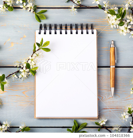 Open paper notebook with empty page and pen on a blue wooden table with cherry blossoms. Top view, flat lay. Open paper notebook with empty page and pen on a blue wooden table with cherry blossoms. Top view, flat lay. 104818366