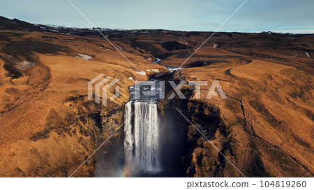 Drone shot of large waterfall in iceland, water stream running down from beautiful brown cliffs. Majestic icelandic skgafoss cascade falling off hills forming spectacular landscape. Slow motion. 104819260