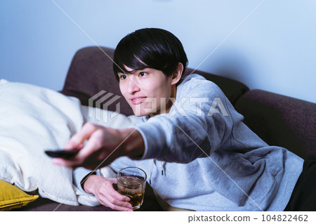 Lazy man in his thirties lying on the sofa and relaxing while drinking alcohol and operating the remote control to change the channel 104822462