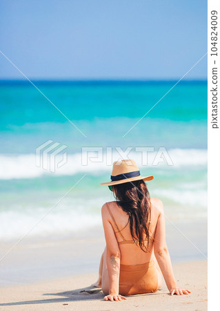 Young beautiful woman in swimsuit and straw hat on the beach vacation sitting on the sand 104824009