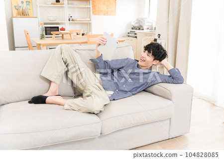 A young man lying on the sofa in the living room and laughing while watching a video on a tablet device A young man lying on the sofa in the living room and laughing while watching a video on a tablet device 104828085