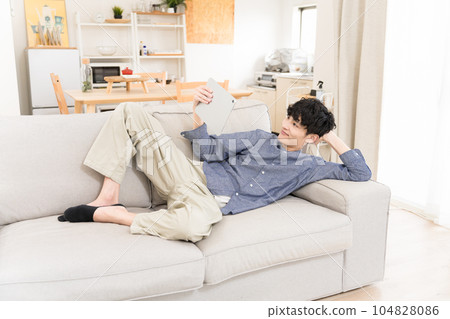 A young man lying on the sofa in the living room and laughing while watching a video on a tablet device A young man lying on the sofa in the living room and laughing while watching a video on a tablet device 104828086