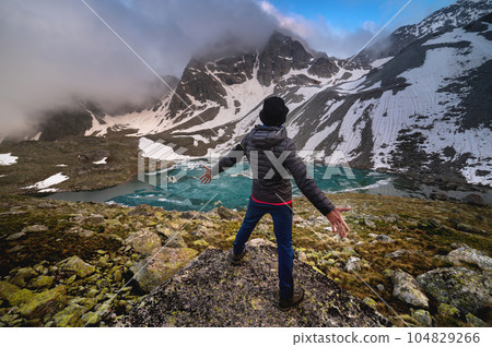 A young man in a downy sweater and panama with his arms outstretched stands in front of a mountain lake in cloudy weather against the backdrop of a clear blue sky through the clouds. A young man in a downy sweater and panama with his arms outstretched stands in front of a mountain lake in cloudy weather against the backdrop of a clear blue sky through the clouds. 104829266