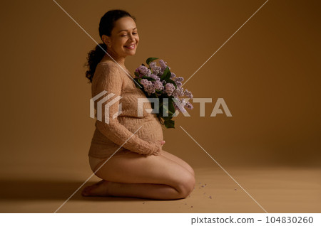 Beautiful positive pregnant woman in elegant clothes, sitting on a beige background with bunch of lilacs, holding belly Beautiful positive pregnant woman in elegant clothes, sitting on a beige background with bunch of lilacs, holding belly 104830260