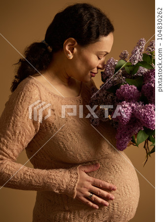 Studio shot of a delightful happy adult pregnant woman with bouquet of lilacs, touching her belly over beige background Studio shot of a delightful happy adult pregnant woman with bouquet of lilacs, touching her belly over beige background 104830262
