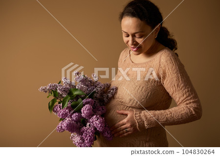 Smiling happy pregnant woman with a bunch of blooming purple lilacs, caressing her belly over beige studio background. Smiling happy pregnant woman with a bunch of blooming purple lilacs, caressing her belly over beige studio background. 104830264