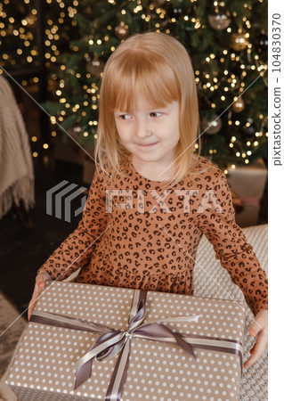 A little blonde girl is sitting on a soft bed in a festive interior decorated in a New Year's style. A child holds a Christmas gift in a craft package. A little blonde girl is sitting on a soft bed in a festive interior decorated in a New Year's style. A child holds a Christmas gift in a craft package. 104830370
