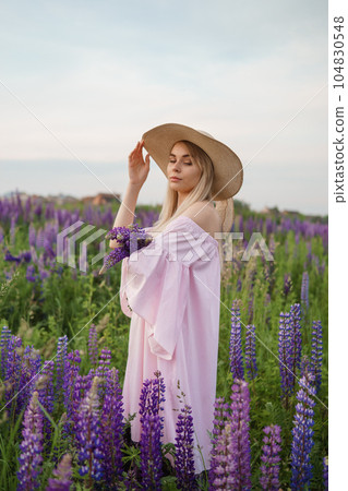 A beautiful woman in a straw hat walks in a field with purple flowers. A walk in nature in the lupin field 104830548