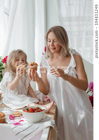 A little blonde girl with her mom on a kitchen countertop decorated with peonies. The concept of the relationship between mother and daughter. Spring atmosphere. 104831249