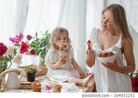 A little blonde girl with her mom on a kitchen countertop decorated with peonies. The concept of the relationship between mother and daughter. Spring atmosphere. A little blonde girl with her mom on a kitchen countertop decorated with peonies. The concept of the relationship between mother and daughter. Spring atmosphere. 104831251