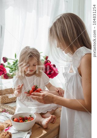 A little blonde girl with her mom on a kitchen countertop decorated with peonies. The concept of the relationship between mother and daughter. Spring atmosphere. A little blonde girl with her mom on a kitchen countertop decorated with peonies. The concept of the relationship between mother and daughter. Spring atmosphere. 104831254