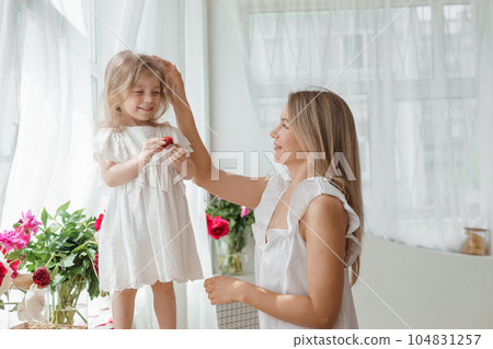 A little blonde girl with her mom on a kitchen countertop decorated with peonies. The concept of the relationship between mother and daughter. Spring atmosphere. A little blonde girl with her mom on a kitchen countertop decorated with peonies. The concept of the relationship between mother and daughter. Spring atmosphere. 104831257