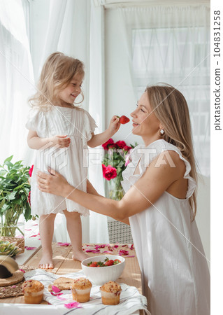 A little blonde girl with her mom on a kitchen countertop decorated with peonies. The concept of the relationship between mother and daughter. Spring atmosphere. A little blonde girl with her mom on a kitchen countertop decorated with peonies. The concept of the relationship between mother and daughter. Spring atmosphere. 104831258