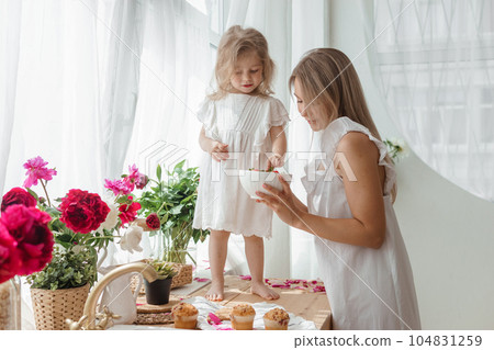 A little blonde girl with her mom on a kitchen countertop decorated with peonies. The concept of the relationship between mother and daughter. Spring atmosphere. A little blonde girl with her mom on a kitchen countertop decorated with peonies. The concept of the relationship between mother and daughter. Spring atmosphere. 104831259